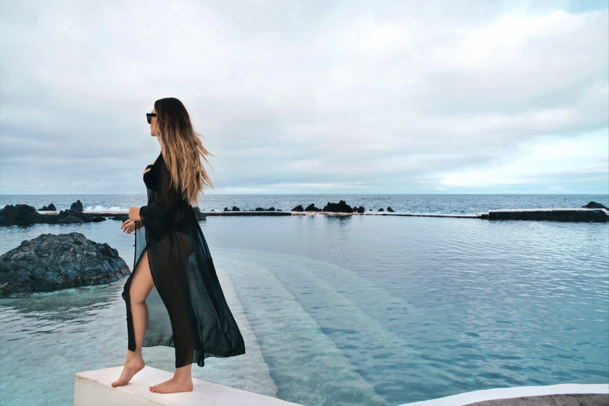 Woman with bikini and black kimono standing in front of natural swimming pools in Madeira