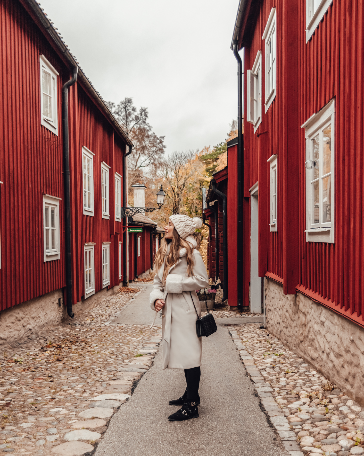 Ida Eriksson i vit fuskpälskappa på en gata bland röda trähus i Wadköping friluftsmuseum, Örebro