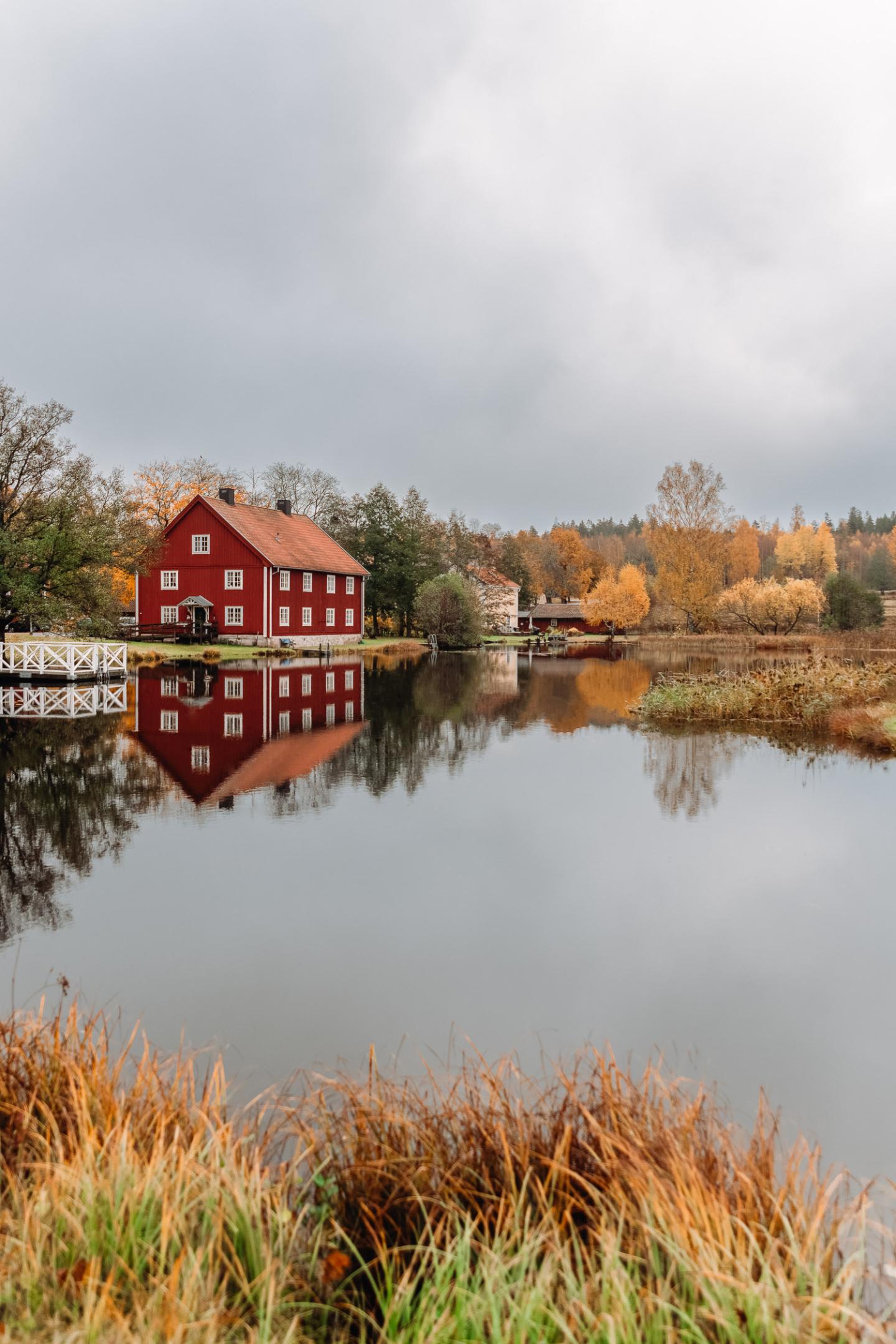 Rött trähus med vita knutar och färgglada höstträd speglas i vattnet i Brevens Bruk, Örebro